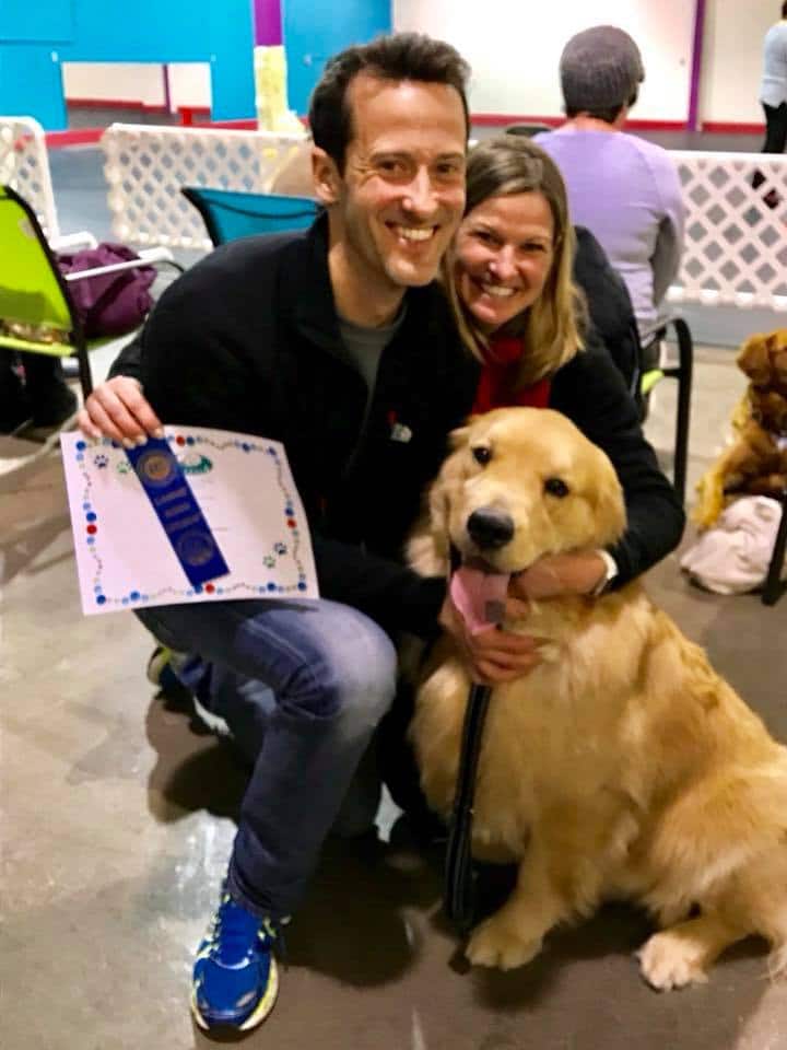 A happy golden retriever named Eddie and his owners proudly show off his graduation diploma and blue ribbon for demonstrating good behavior at adult dog training classes at Augusta Dog Training.